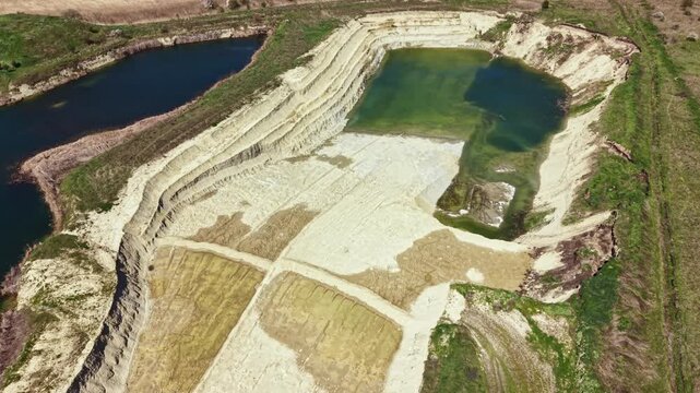 A sand quarry has multiple water pools with clear blue and green hues. The quarry shows different layers of earth from mining activity in bright daylight.