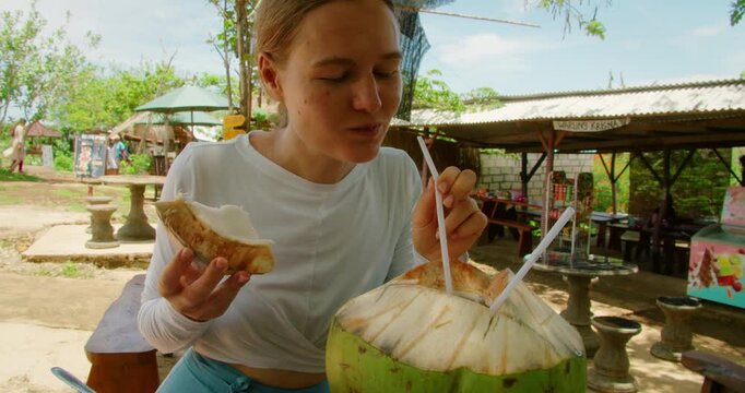 Woman drinks fresh coconut water through straw while eating fruit slice outdoor rustic cafe in Bali