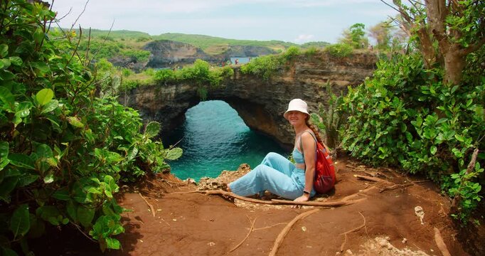Woman tourist with backpack sits on cliff edge overlooking Broken Beach arch Nusa Penida Bali Indonesia