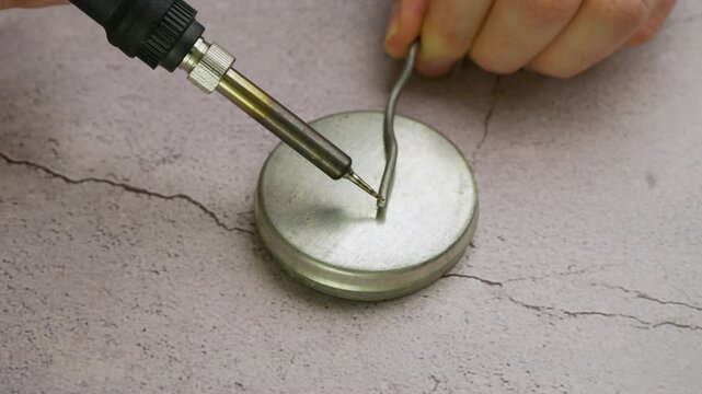 Detailed shot of a person using a soldering iron to melt solder wire onto a round metal jar lid on a grey workbench surface