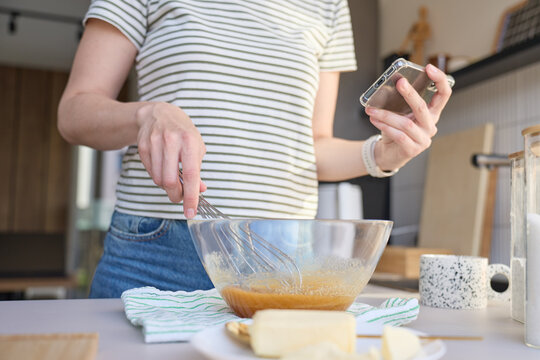 Woman whisking batter in glass bowl while holding smartphone in kitchen