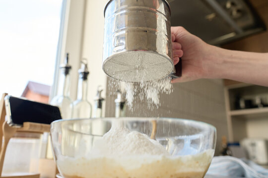 Hand sifting flour through metal sieve into glass bowl in kitchen