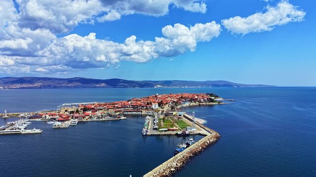 Nesebar Old Town sits by the Black Sea with boats docked by the shore. Buildings with red roofs are visible under a blue sky with clouds. The waterfront area is busy during daylight.