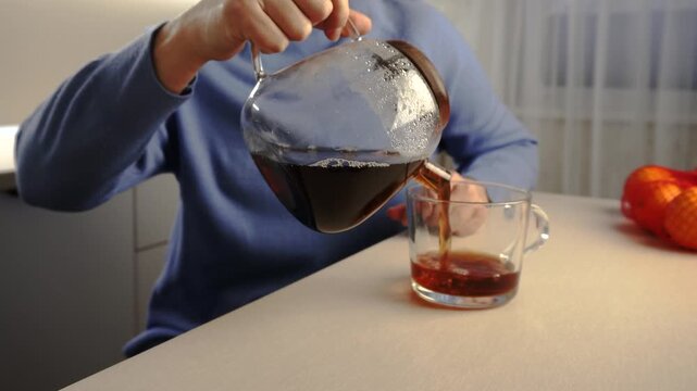 A man pours dark coffee from a glass coffee pot into a clear glass cup. The man wears a blue sweater while the background features a soft light and red oranges.