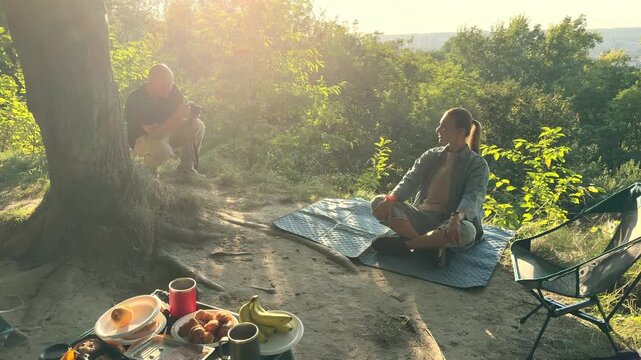 Man photographer taking pictures of a woman sitting on a picnic blanket on a hill. Behind the scenes of a photo shoot in nature with a scenic city view at sunset. Concept of content creation 