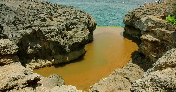 Natural tidal pool surrounded by massive grey rocks with people in distance enjoying view at Angel Billabong beach Nusa Penida