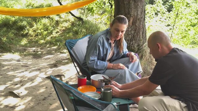 Close-up of campers preparing a hot meal using a portable gas stove and a metal pot. Man and woman cooking outdoors during a hiking trip. Concept of outdoor survival and camping gear