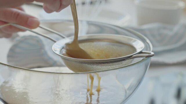 Hand straining sauce through metal sieve into glass bowl. Concept of home cooking and baking preparation