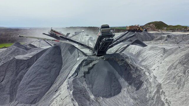 Heavy machinery works in a granite quarry to extract stone while dust rises from the ground and clouds cover the sky.