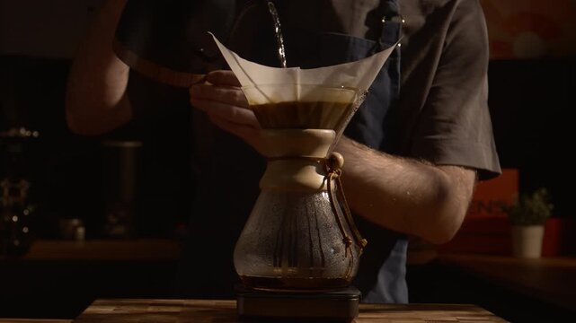 Man preparing filter coffee with Chemex, pouring water in slow motion, dramatic shadows and sunlight, aesthetic specialty coffee process.
