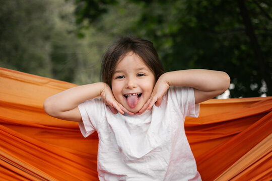 Young girl laughs with tongue out in bright orange hammock surrounded by green trees outdoors