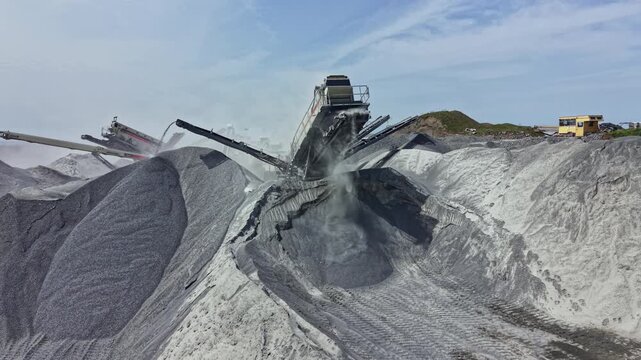 Large equipment works in granite quarry to extract stone and move materials around site as dust and debris fill the air