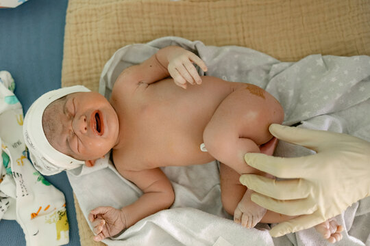 Newborn baby cries softly while wrapped in blanket as gloved hand gently touches leg in hospital room