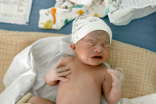 Newborn baby cries softly while wrapped in blanket on hospital bed surrounded by medical papers and soft textiles