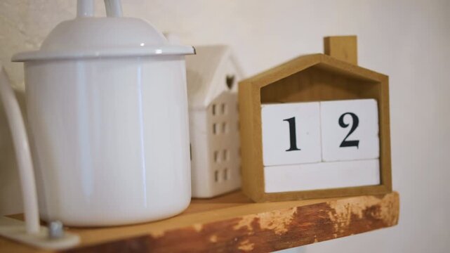 Wooden house calendar on kitchen shelf, white enamel pot beside numbered block, terracotta jug in background, morning prep routine as home cook checks date before baking, rustic display with soft