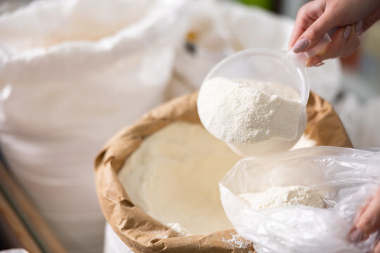 Womens hands fill single-use plastic bag with white flour. In self-service store, customer pours flour from bag and disposable container, she uses ladle to collect white flour in sack..