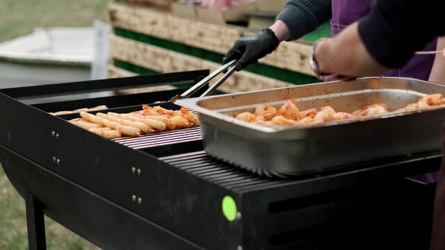 Cook using metal tongs to flip shrimp on a charcoal grill. Chef preparing grilled seafood skewers outdoors. Appetizing appetizer cooking process for dinner.