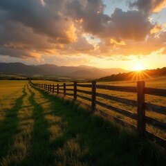 Obraz premium Wooden fence divides green grass pasture at sunset. Sun rays shine over rolling hills and distant mountains. Dramatic clouds fill the golden sky over farmland.