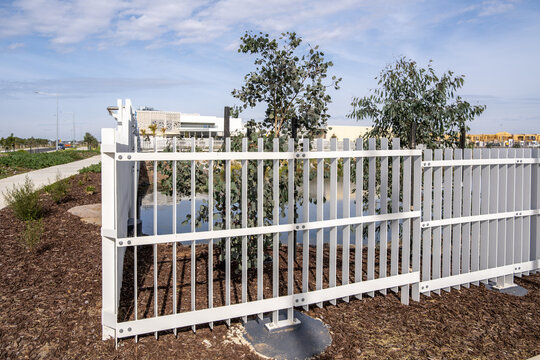 White metal pool safety fence beside a stormwater retention pond in a new suburban Melbourne housing estate, Tarneit, Australia. Young eucalyptus trees and mulched garden beds line the water's edge