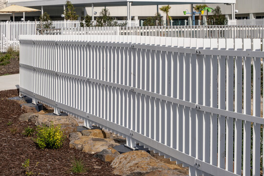 A long white metal palisade fence runs diagonally through the frame alongside a landscaped garden bed. Contemporary barriers or fencing in public open space or recreational facility