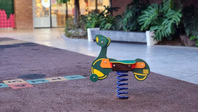A colorful green and yellow motorcycle-shaped spring rider on a rubberized safety surface at a modern outdoor children's playground.