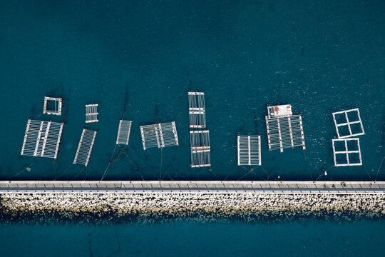 Aerial view of floating oyster farm structures and rectangular cages in the deep blue sea along a stone breakwater at Itoshima Peninsula Itoshima, Fukuoka, Japan.
