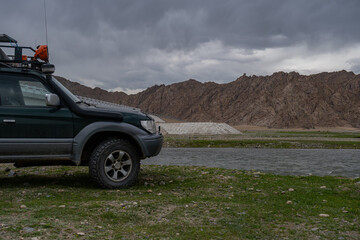 Fototapeta premium Off-road 4x4 vehicle with expedition roof rack parked near a mountain river. Adventure travel and overlanding concept in wild rocky landscape under cloudy sky.