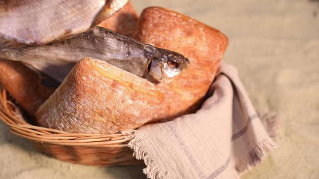 Woman taking dried fish from wicker basket with loaves of bread on sand, closeup