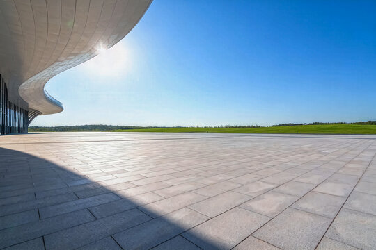 A bright sunny open square paved with stone tiles, featuring a curved modern building facade, wide green lawn under clear blue sky.