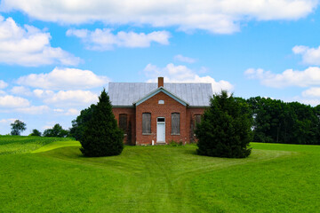Fototapeta premium An old brick house with boarded windows sits between two large bushes on a green grassy hill under a blue sky with clouds surrounded by trees in the countryside