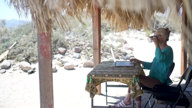 A mature woman types on her computer under a palapa at a beach in Baja California, Mexico while enjoying the warm sunshine.