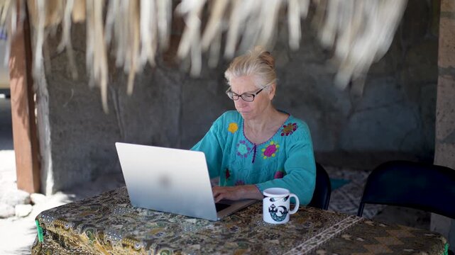 Mature woman is focused on her computer while working under a palapa on a sunny tropical beach in Baja California Mexico.