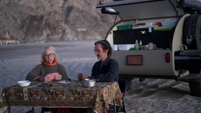 A mature man and woman enjoy a card game by their teardrop camper trailer on a beach in Baja California Mexico at sunset.