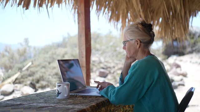 Mature woman focuses on computer while seated under palapa on beach in Baja California. She enjoys nature while working remotely.