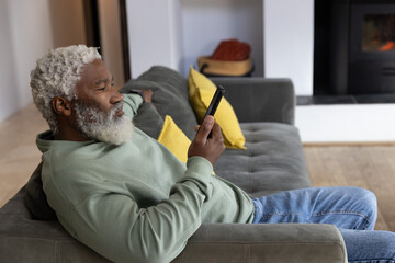 Naklejka premium Senior African American man reclining on gray sofa in living room, holding phone near fireplace