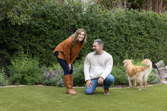 Couple in knit sweaters kneeling and laughing in backyard on manicured lawn near bench and dog
