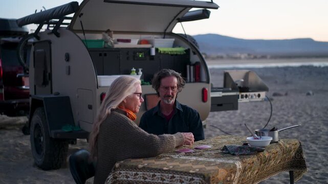 A mature man and woman enjoy card games next to their teardrop camper on the beach during sunset in Baja California, Mexico.