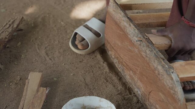 Closeup black carpenter hammering plank, focused strikes into timber, sawdust cloud, hand bracing board, gritty sand floor, red bracelet, outdoor workshop atmosphere, traditional joinery motion