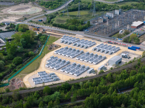 Aerial view of Battery Energy Storage Systems with rows of white storage containers, electrical substation, and surrounding green trees in Knottingley, England, United Kingdom.