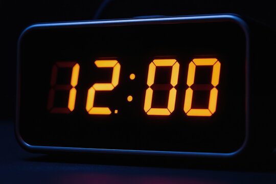 Digital clock shows 12 00 with bright orange illuminated digits. Device display shines in dark room. Modern electronic timekeeper shows hours and minutes.