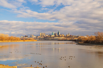 Naklejka premium A city skyline rises behind a wide river under a cloudy sky.