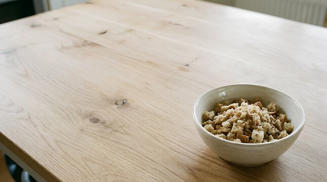 Small ceramic bowl filled with stale bread crumbs placed in the bottom‑right corner of a large light wooden table, most of the table surface kept empty as negative space.