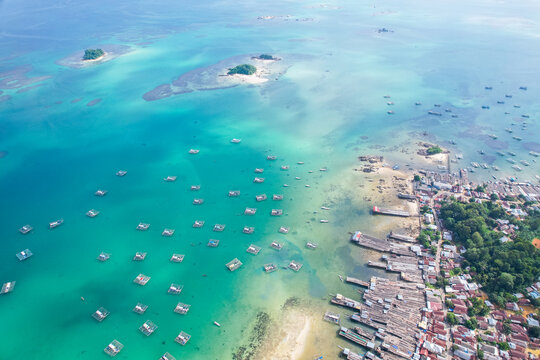 High-angle aerial view of a traditional fishing village in Tanjung Binga, Belitung, Indonesia, featuring coastal houses, fish drying platforms, and boats on turquoise water.