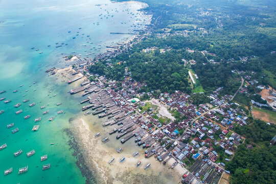 High-angle aerial view of a traditional fishing village in Tanjung Binga, Belitung, Indonesia, featuring coastal houses, fish drying platforms, and boats on turquoise water.