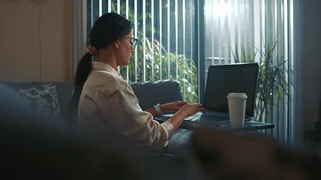 Woman wearing glasses typing on laptop at home by window with coffee creating calm productive remote work atmosphere in natural daylight using modern technology