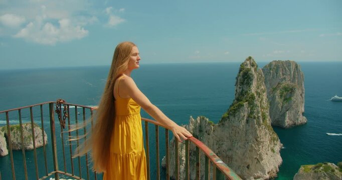 Woman in sundress stands on Capri cliff balcony facing Faraglioni rocks