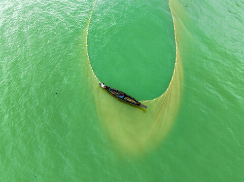 Aerial view of a fisherman in a small wooden boat casting a large golden fishing net in the green water of Rangamati, Chittagong Division, Bangladesh.