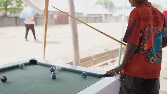 Young black man lining pool shot under canopy with colorful shirt, outdoor table, cue tip near ball, focused stance, sunlight on sand, community background, casual competition atmosphere