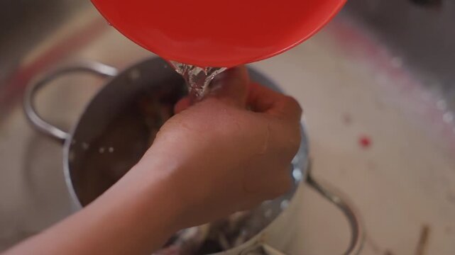 Pouring water over strainer in sink with red bowl, hands steady and controlled as unspecified helper rinses kitchenware droplets and suds, metal drain and bowl, domestic rinse routine for meal prep