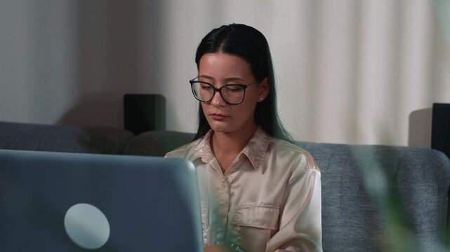 Woman wearing glasses focused working on laptop at home on sofa creating attentive productive remote work atmosphere in calm home environment using modern technology
